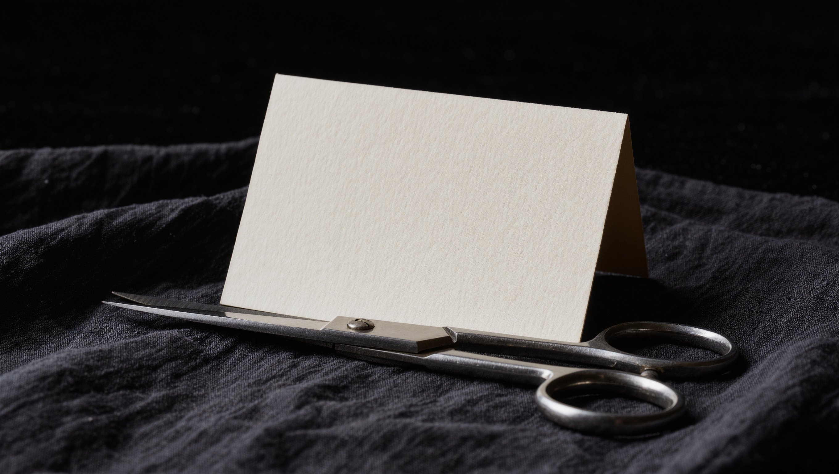 Aufgestellte matte Karte ohne Aufdruck neben einer Schere auf dunklem Leinen im weichen Seitenlicht
